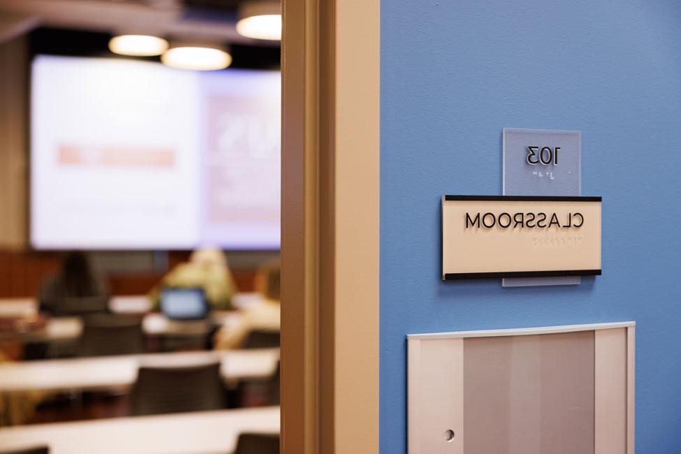 Exterior of entry to classroom. Classroom wall sign is in the foreground with CED students sitting in a classroom in background.